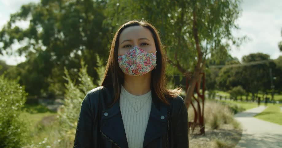 A woman wearing a floral pattern medical mask raises her head to look into the camera while standing on a walkway in a green space in a park