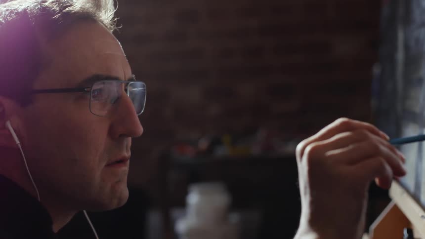 A man wearing eyeglasses and listening to something on his earbuds is painting on a canvas on an easel in front of him