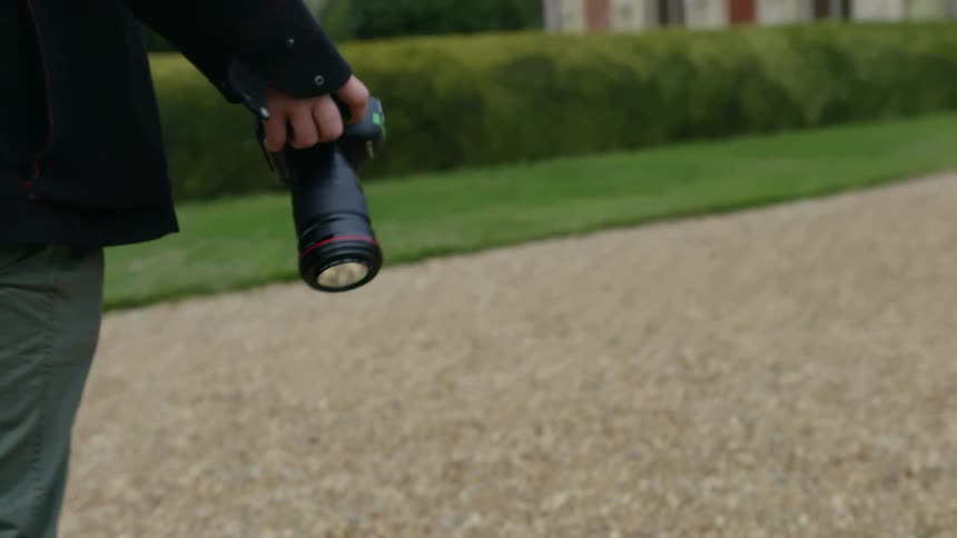A person walks on a path towards a building while they hold a camera in one hand