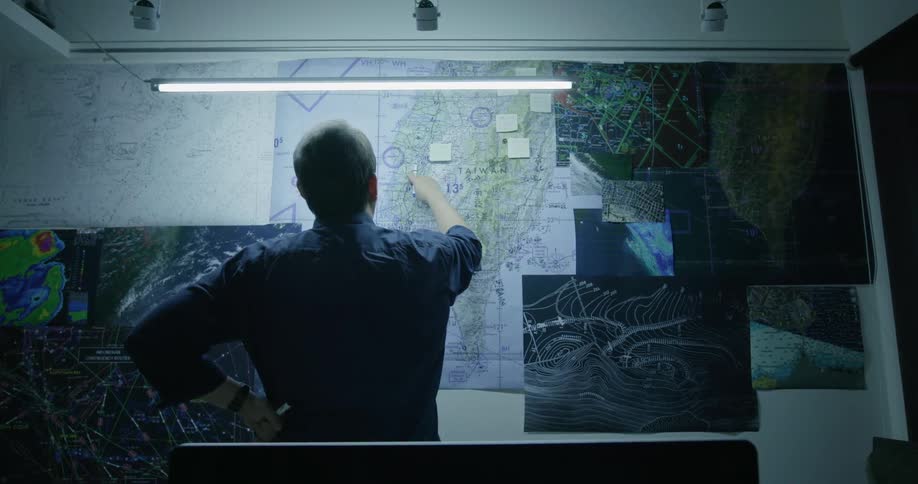 A man stands in a room with several different types of maps on the wall as he points to a location on one of them and studies it more closely