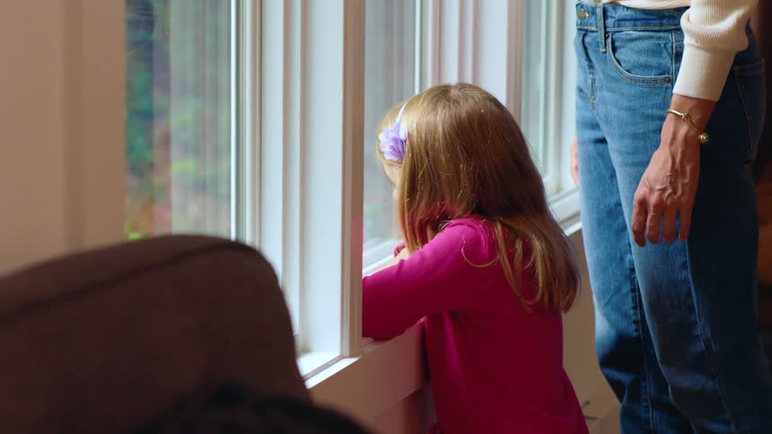 A woman walks over to a child leaning against a window looking out and comforts her by caressing her hair
