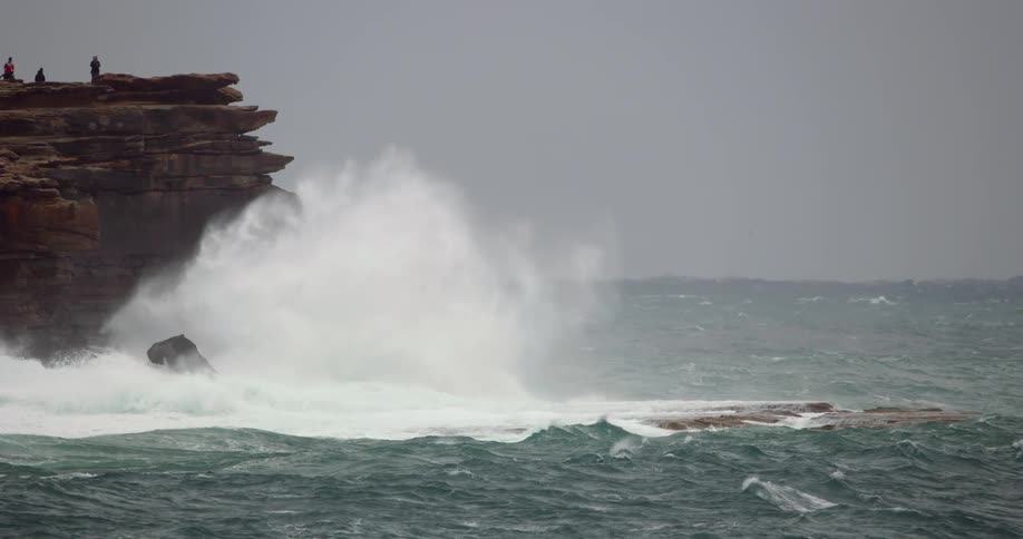 Ocean waves crash into a rocky cliff where a small group of people stand on a foggy day