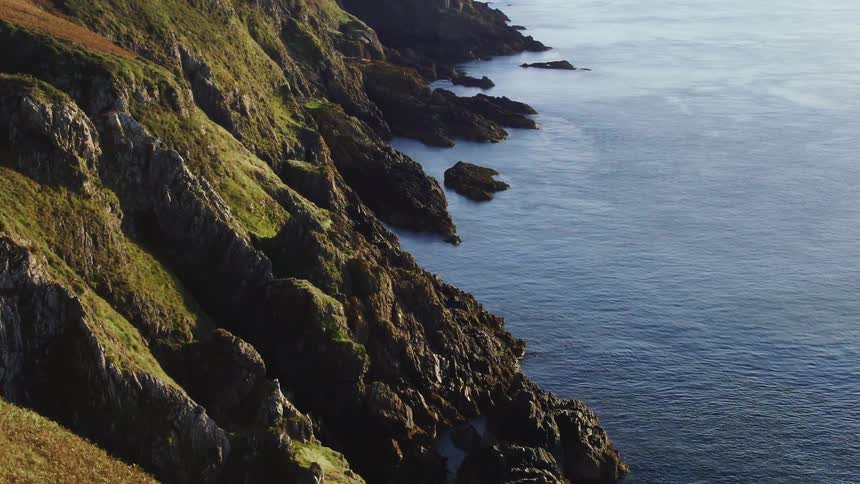 Aerial view of area of rocks with a very steep side at the edge of the calm sea and cliff road