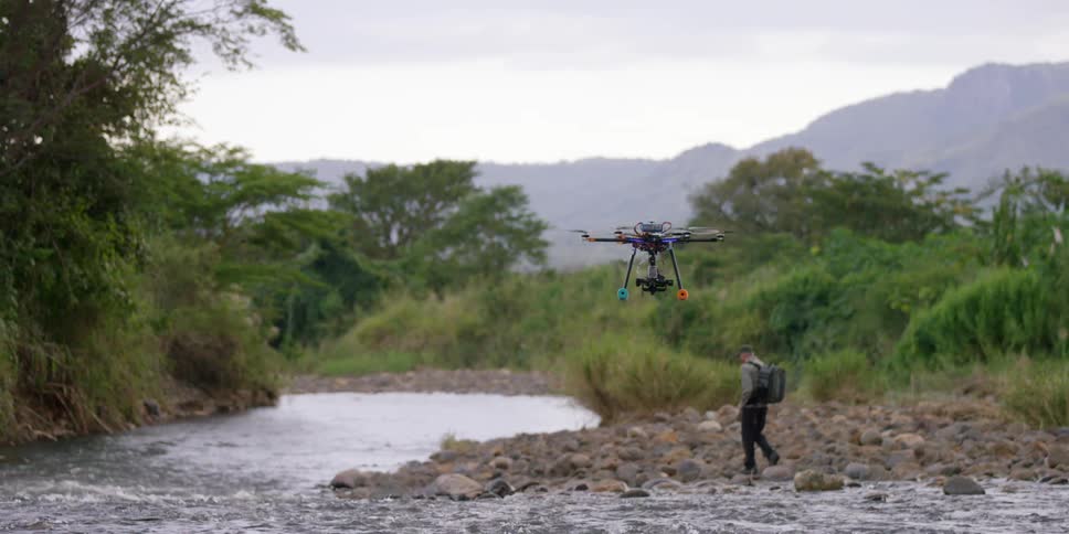 A drone following a man who is walking along a rocky river bank