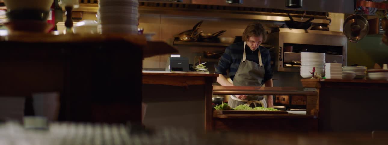 A man in a kitchen wipes off his knife and scrapes vegetables on a cutting board