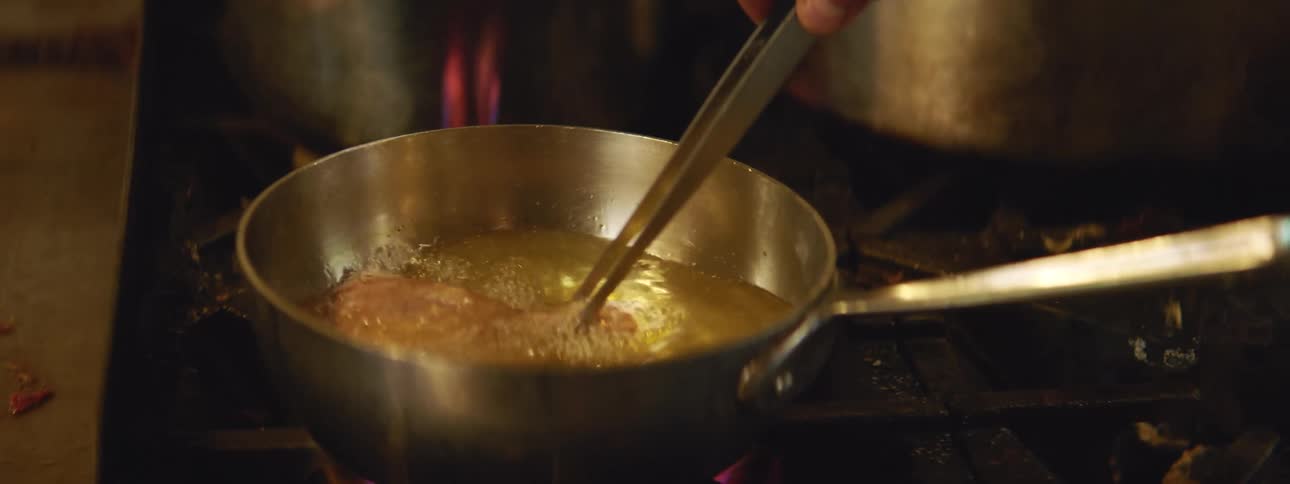 Food being boiled in oil in a saute pan on a stove top with a pair of tongs