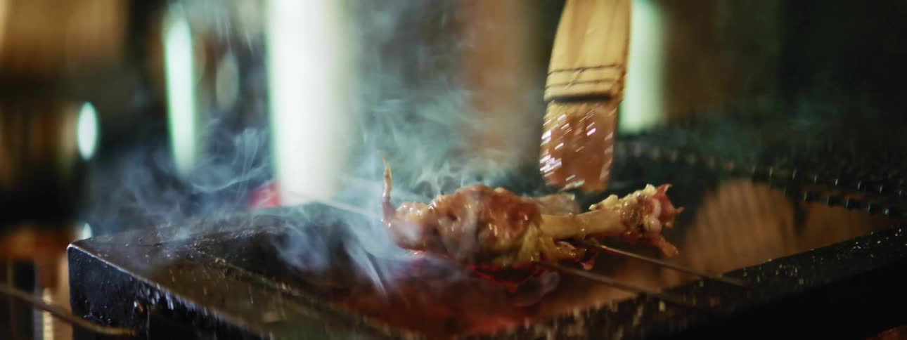 Meat being grilled on a pair of skewers while being glazed with a pastry brush