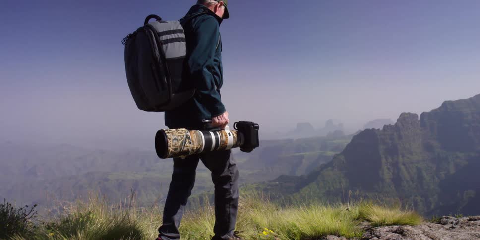 A man in jeans carries a camera on a rocky path