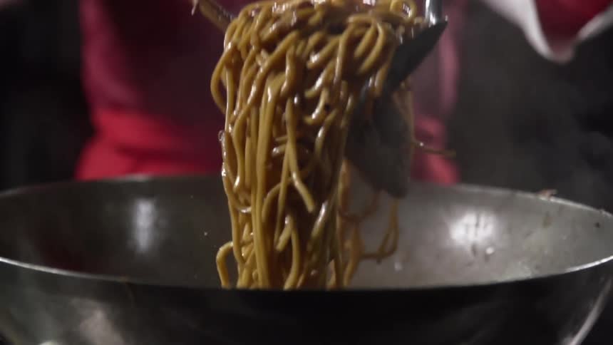 A cook uses a spatula and a pair of tongs to lift noodles from a steaming wok before letting the noodles fall back inside
