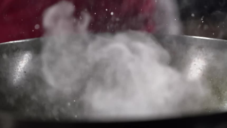 Oil drops pop up and smoke and steam rise from a wok