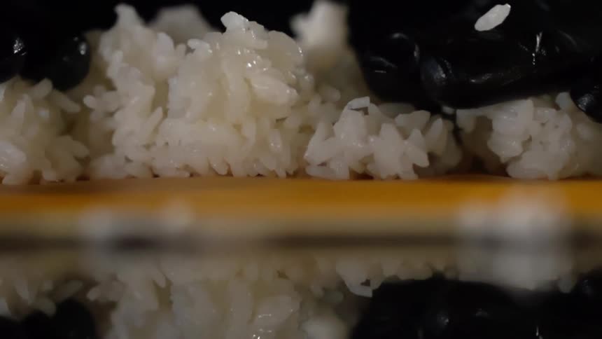 Gloved hands spread sushi rice out on a mat on a black mirror surface