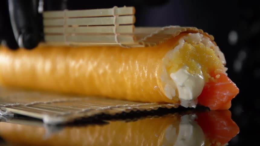Chef wearing black latex gloves rolls sushi with a bamboo mat over a reflective surface
