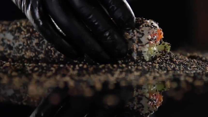 A sushi roll being twisted in a pile of sesame seeds by a pair of hands covered in black gloves