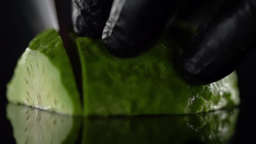 A person wearing black disposable gloves slices through an avocado half with a knife