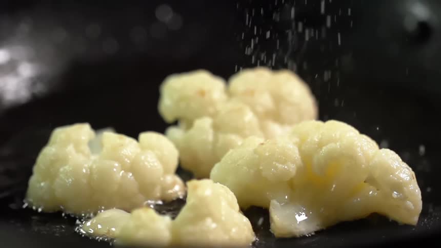 Salt sprinkles on pieces of cauliflower being cooked in a pan