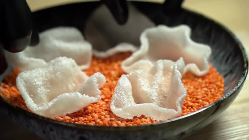 Black gloved hands place rice chips into a bowl of lentils