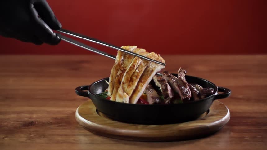 Sliced chicken is placed next to steak on top of cooked peppers in a cast iron serving pan