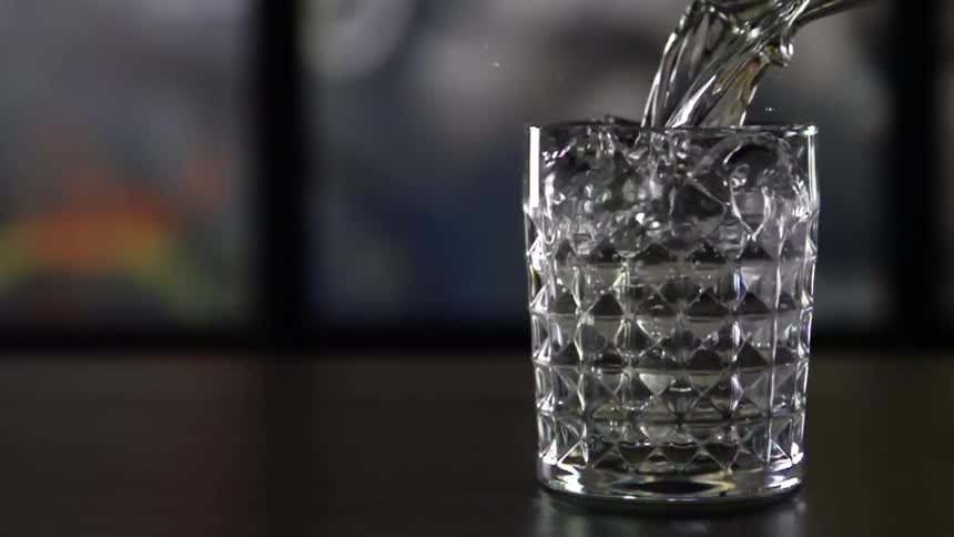 An alcoholic beverage is poured into a clear glass overflowing it and causing some to splash out