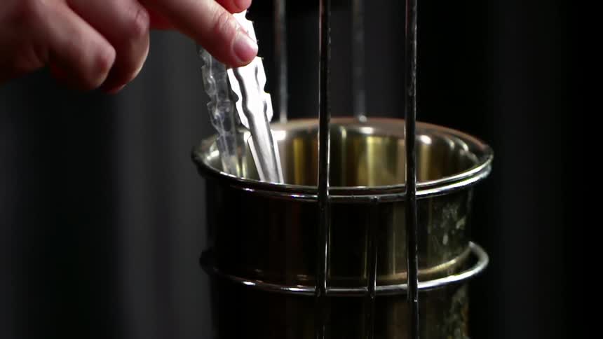A person uses small tongs to pick a charcoal cube up out of a metal bowl