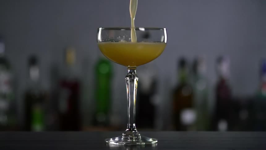 A yellowish cocktail is being poured into a glass in a bar