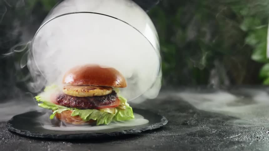 A lid is taken off of a presentation plate revealing a burger and cloud of dry ice smoke