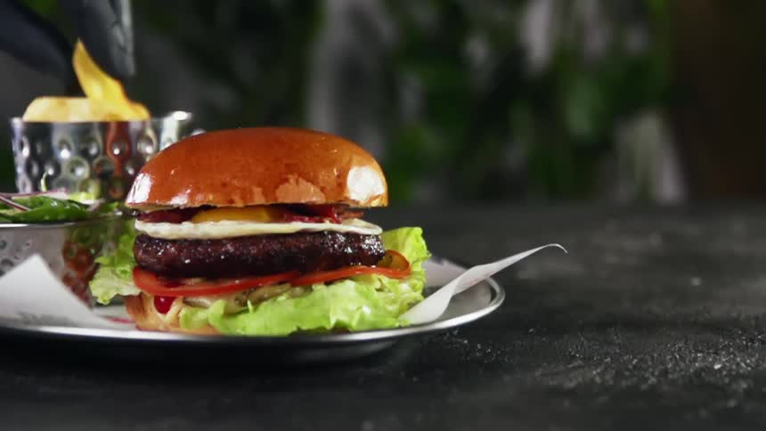 A burger with a fried egg and bacon and a person with black gloves taking fries in the background