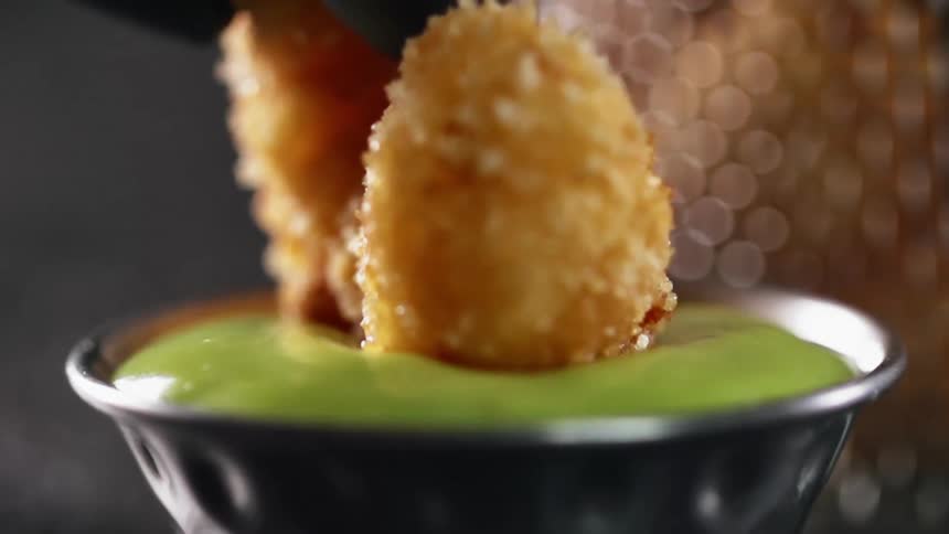 A breaded shrimp being dipped into guacamole in a tin dipping bowl