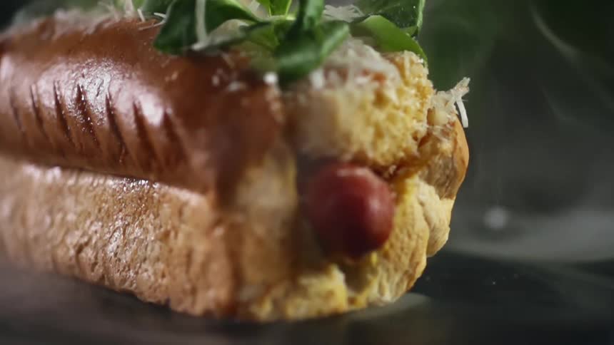 The end of a hot dog sticks out from a bun with gourmet toppings and fresh herbs