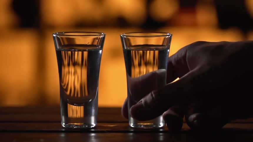 Two tall shot glasses are located on top of a bar counter and one is picked up and taken away