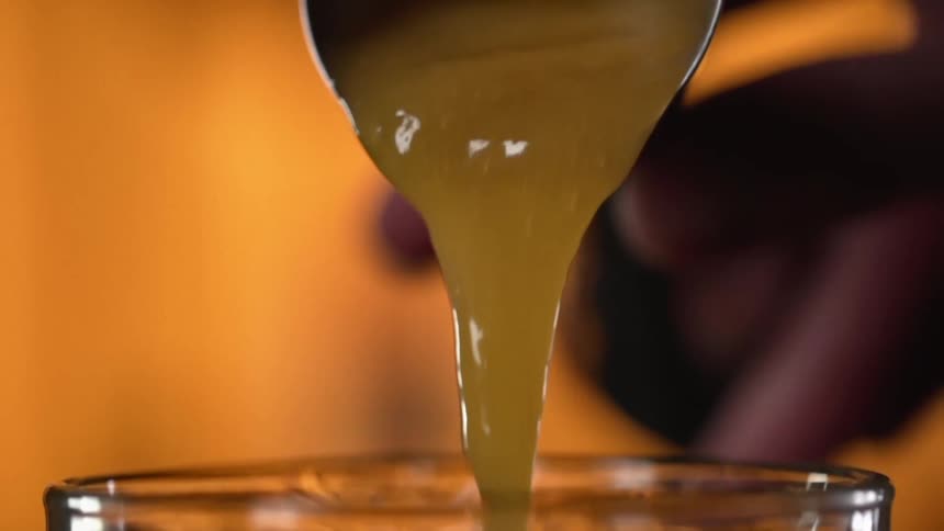 A person is pouring a thick sauce from a small metal cup into a clear glass container