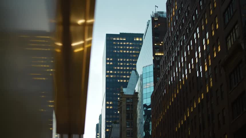 Window lights in city buildings glow at dusk while a glass facade reflects sky and city