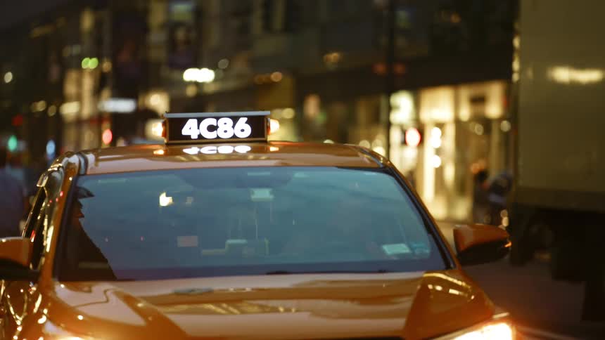 A taxi sitting on a busy city street as other cars drive by and people walk on the street