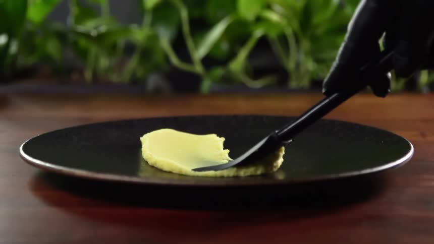 Chef wearing black latex gloves smears yellow cream with a spatula over a black ceramic dish