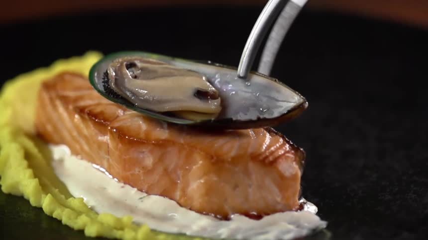A pair of tongs is used to place a muscle on top of a piece of seafood and sauce