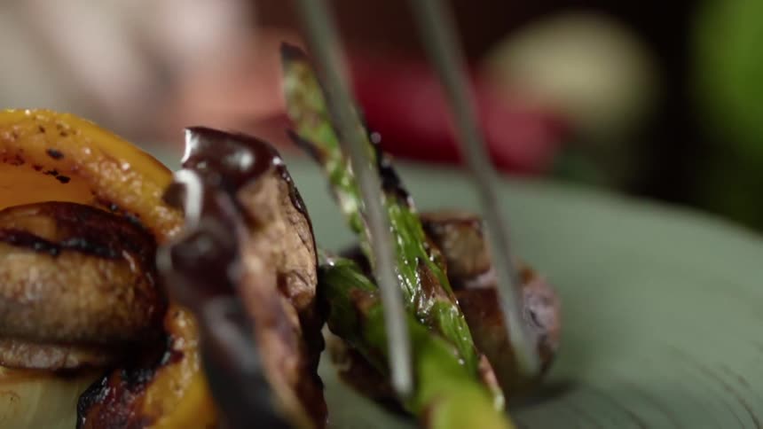 A piece of roasted asparagus is gently placed on top of other vegetables on a green plate