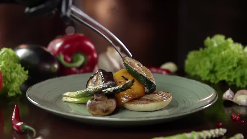 A person uses a pair of tongs to place some grilled vegetables on a plate