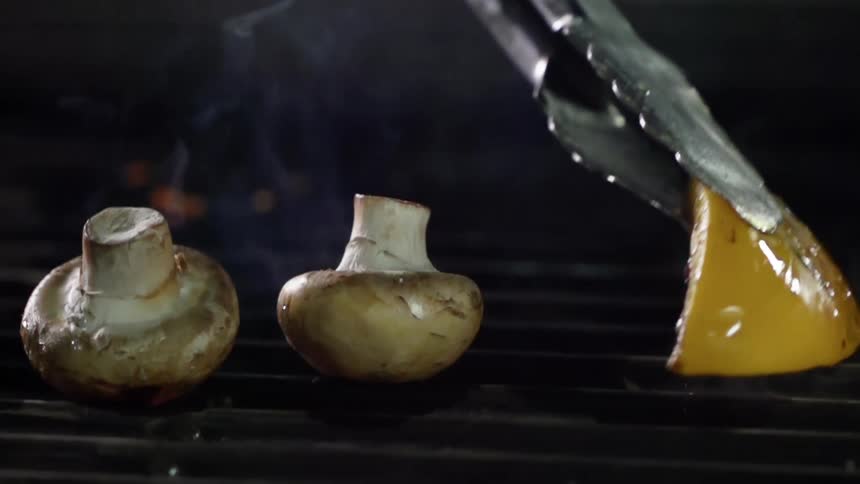 A pair of tongs picks up a piece of potato cooking on a grill beside mushrooms
