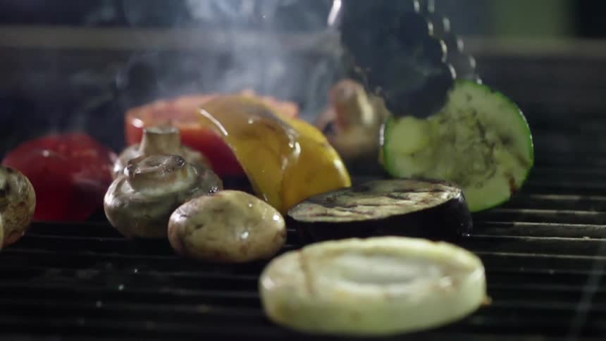 A pair of metal tongs flips over a vegetable slice on a grill
