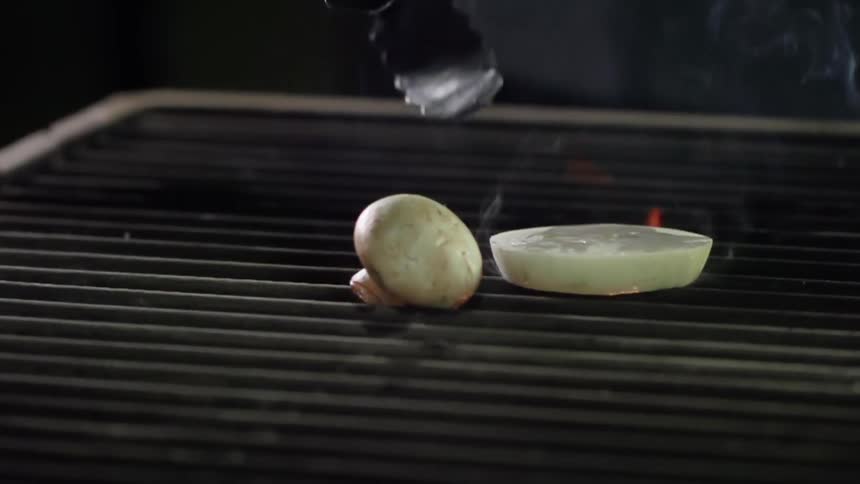 Tongs place onion slice onto flaming smoking grill next to button mushroom