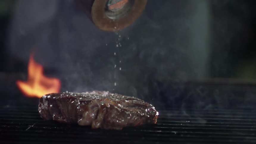 Salt is sprinkled atop a steak being cooked on an open flame grill