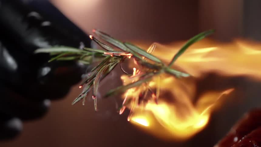 A person wearing a dark glove holds a piece of rosemary while using a torch to singe it
