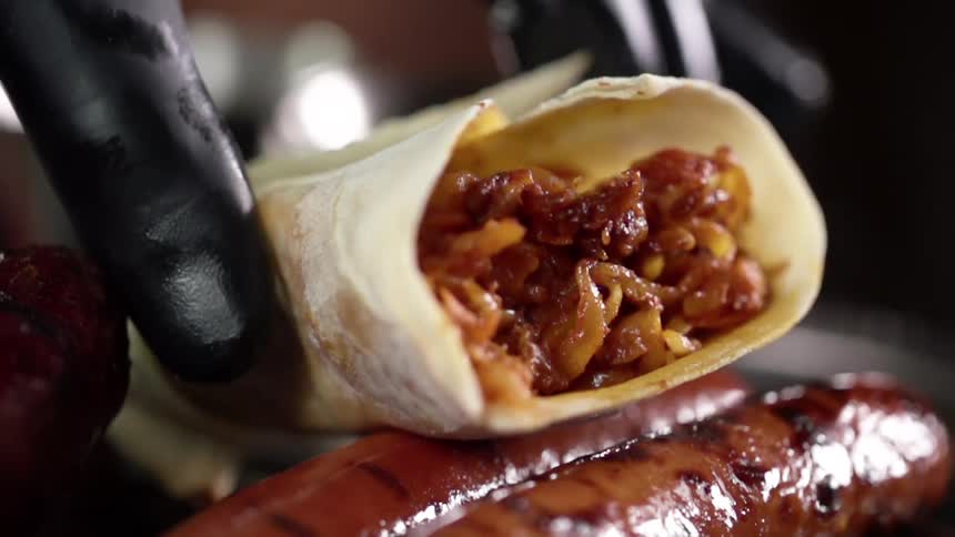 Chef wearing black latex gloves places flour tortilla burrito over grilled sausage