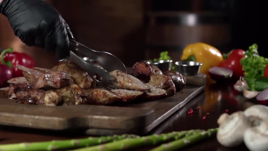 Chef wearing black latex gloves places cooked meats over a wooden board near vegetables
