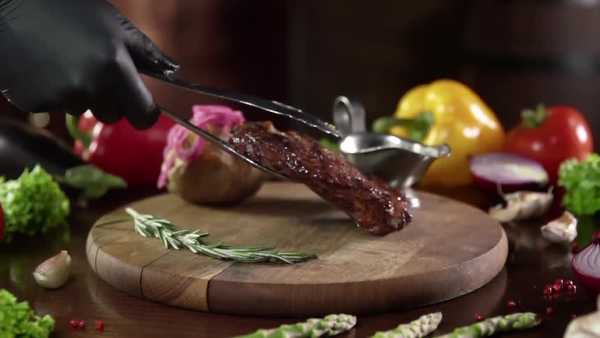 Grilled steak is placed on a round wooden cutting board surrounded by vegetables