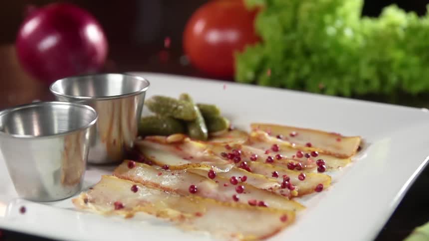 Pink peppercorns are dropped onto a plate with thinly sliced pork belly