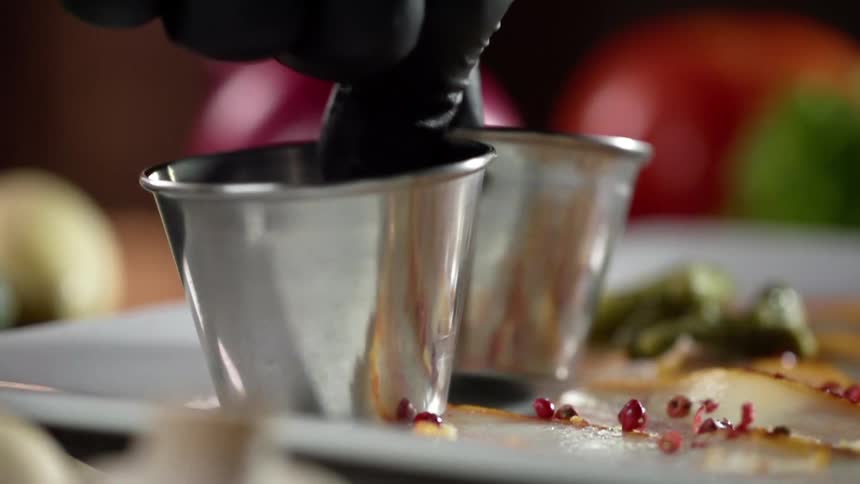 A person wearing black disposable gloves places two small metal cups onto a plate with food on it