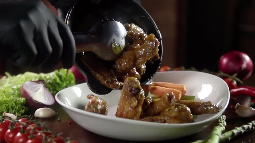 A person is scooping sauce covered pieces of cooked chicken onto a glass dish