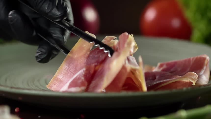 A gloved hand places cured meats on a platter using tongs