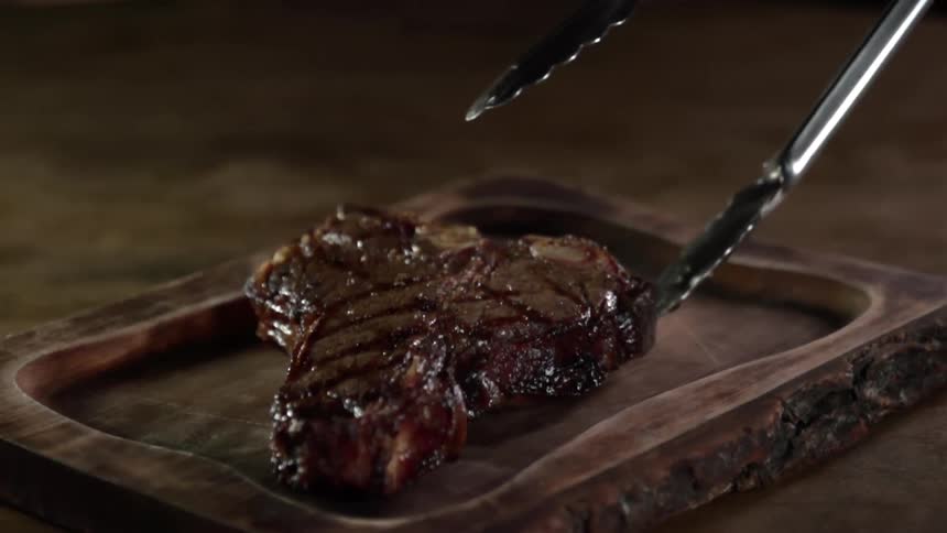 A large piece of grilled steak is placed on a wooden tray using a pair of metal tongs