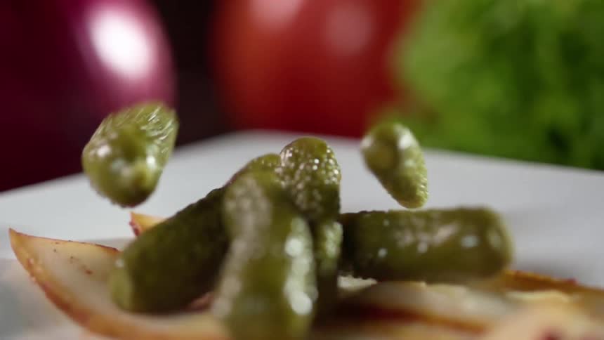 Bunch of pickles falling over bacon slices in a white ceramic plate near produce